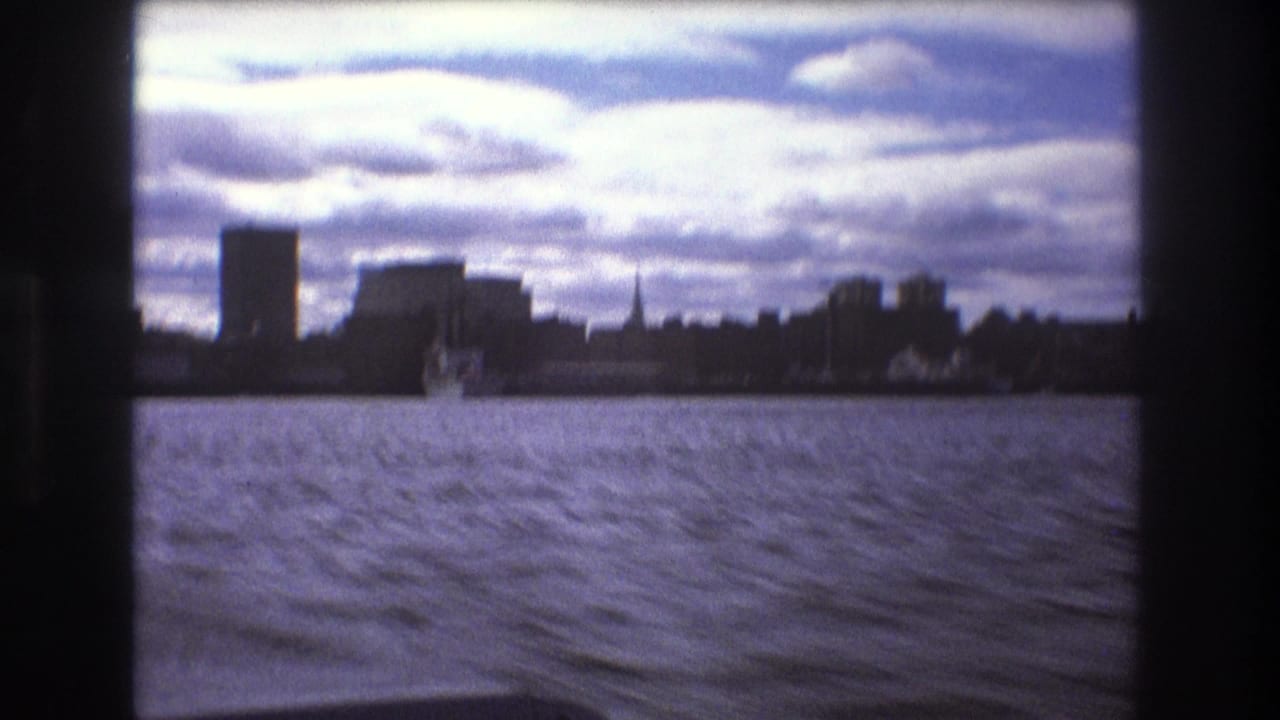 Boston, 1975, with choppy bay water, a small distant boat, low silhouetted waterfront buildings, and bright layered clouds seen from a moving vessel.