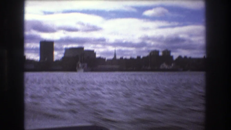 Boston, 1975, with choppy bay water, a small distant boat, low silhouetted waterfront buildings, and bright layered clouds seen from a moving vessel.