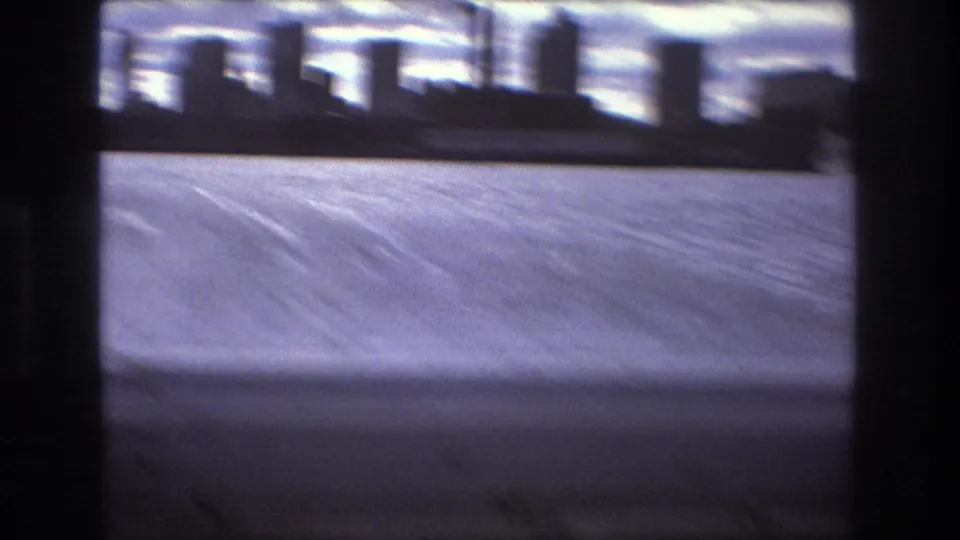 Blurred 1975 Boston waterfront view from a fast-moving boat, with choppy water, distant buildings, cloudy sky, and dark 8mm frame edges.