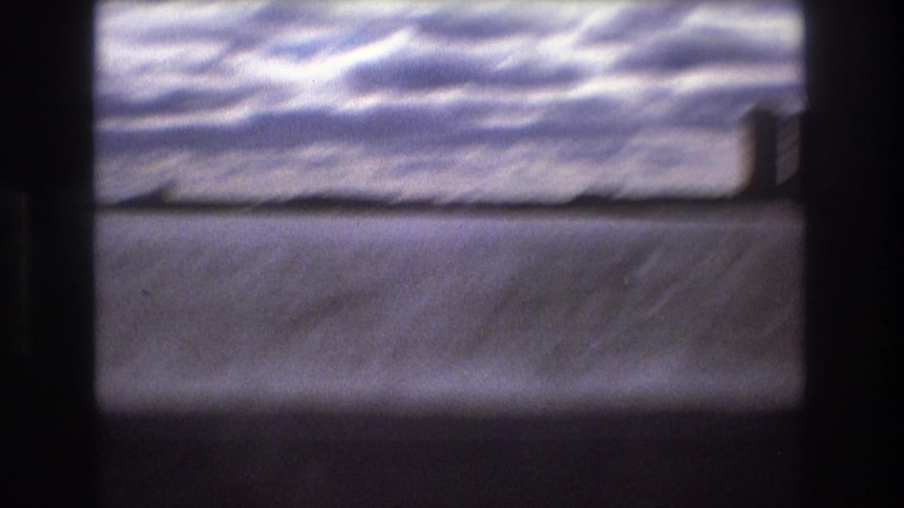 Soft, blurred 1975 Boston harbor view with low shoreline forms beyond water and a clouded sky, framed by dark 8mm gate edges.