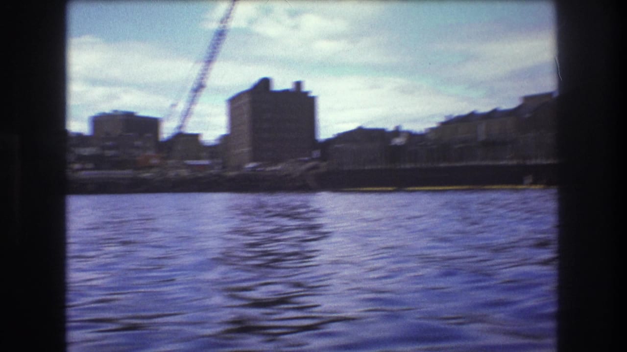 1975 Boston waterfront from a moving boat, showing rippled harbor water, blocky buildings, a construction crane, and a pier-like edge under partly cloudy sky.