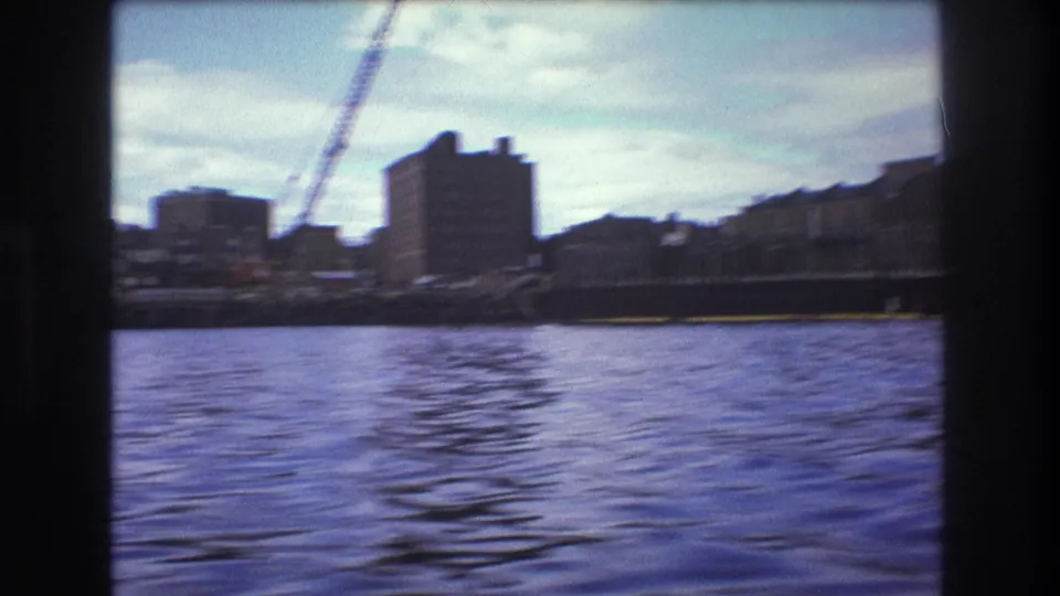 1975 Boston waterfront from a moving boat, showing rippled harbor water, blocky buildings, a construction crane, and a pier-like edge under partly cloudy sky.