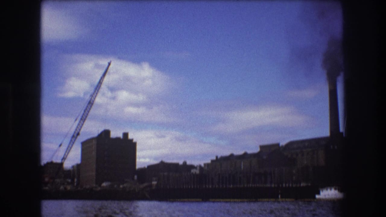 1975 Boston 8mm frame showing an industrial riverside building, a construction crane, and smokestacks across the water under a blue cloudy sky.