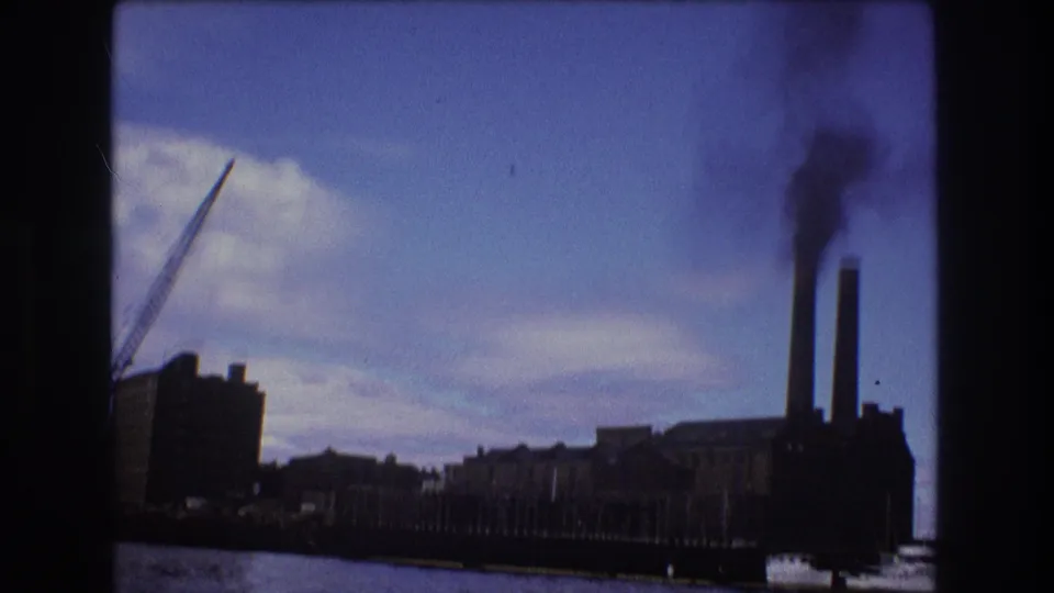 1975 Boston 8mm frame of factory-like waterfront buildings, twin smokestacks emitting dark smoke, and a crane beside the river.