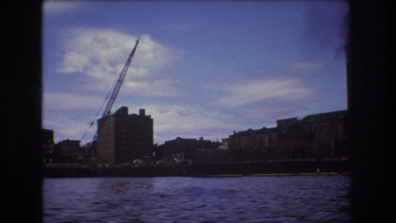 1975 Boston 8mm frame showing a crane over waterfront industrial buildings, with rippled river water and soft home-movie color.