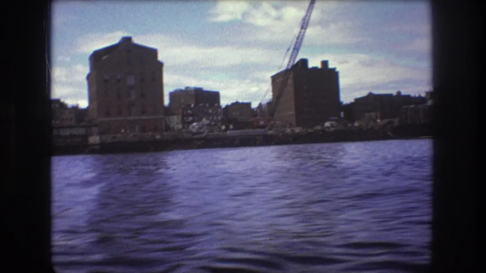 1975 Boston 8mm frame with rippling water in the foreground and industrial waterfront buildings plus a crane in the distance.