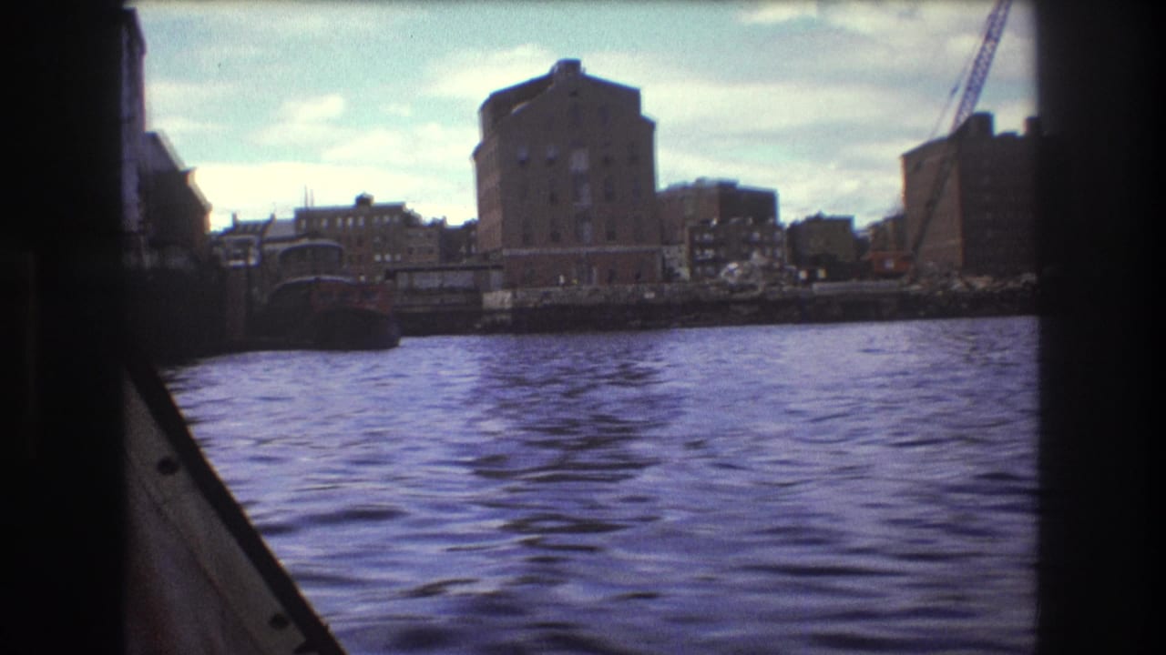 1975 Boston waterfront view from a moving boat, with rippled harbor water, shoreline buildings, and a crane under a partly cloudy sky.