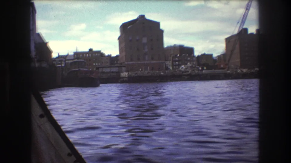 1975 Boston waterfront view from a moving boat, with rippled harbor water, shoreline buildings, and a crane under a partly cloudy sky.