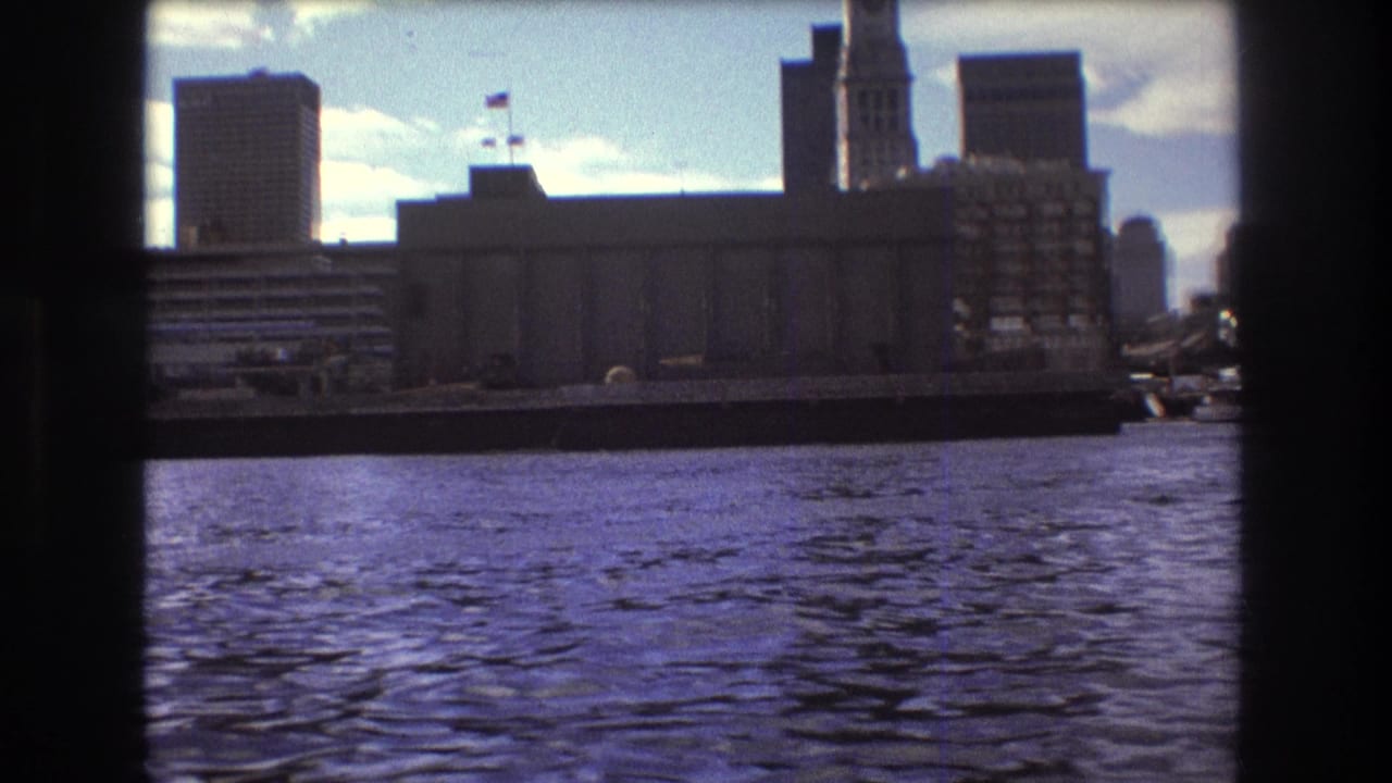 1975 Boston view from the bay, with a large waterfront structure, tall city buildings, a flag, and rippled water in the foreground.