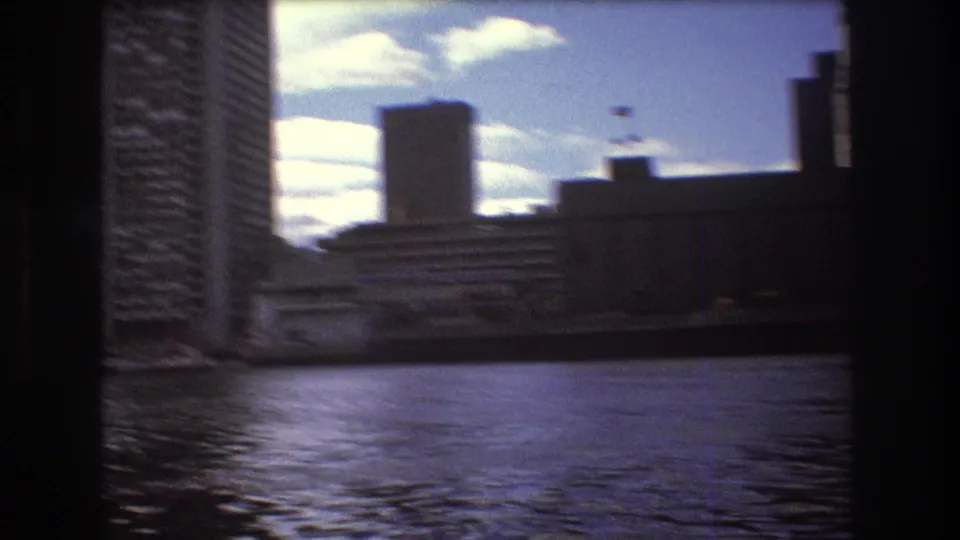 Soft 1975 Boston boat-view frame with rippled bay water, high-rise buildings, waterfront structures, and dark film borders.