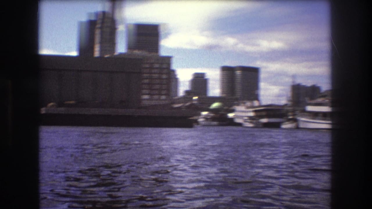 1975 Boston harbor view from a moving boat, showing rippling water, docked vessels, waterfront buildings, and taller office blocks in the background.