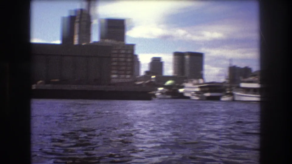 1975 Boston harbor view from a moving boat, showing rippling water, docked vessels, waterfront buildings, and taller office blocks in the background.