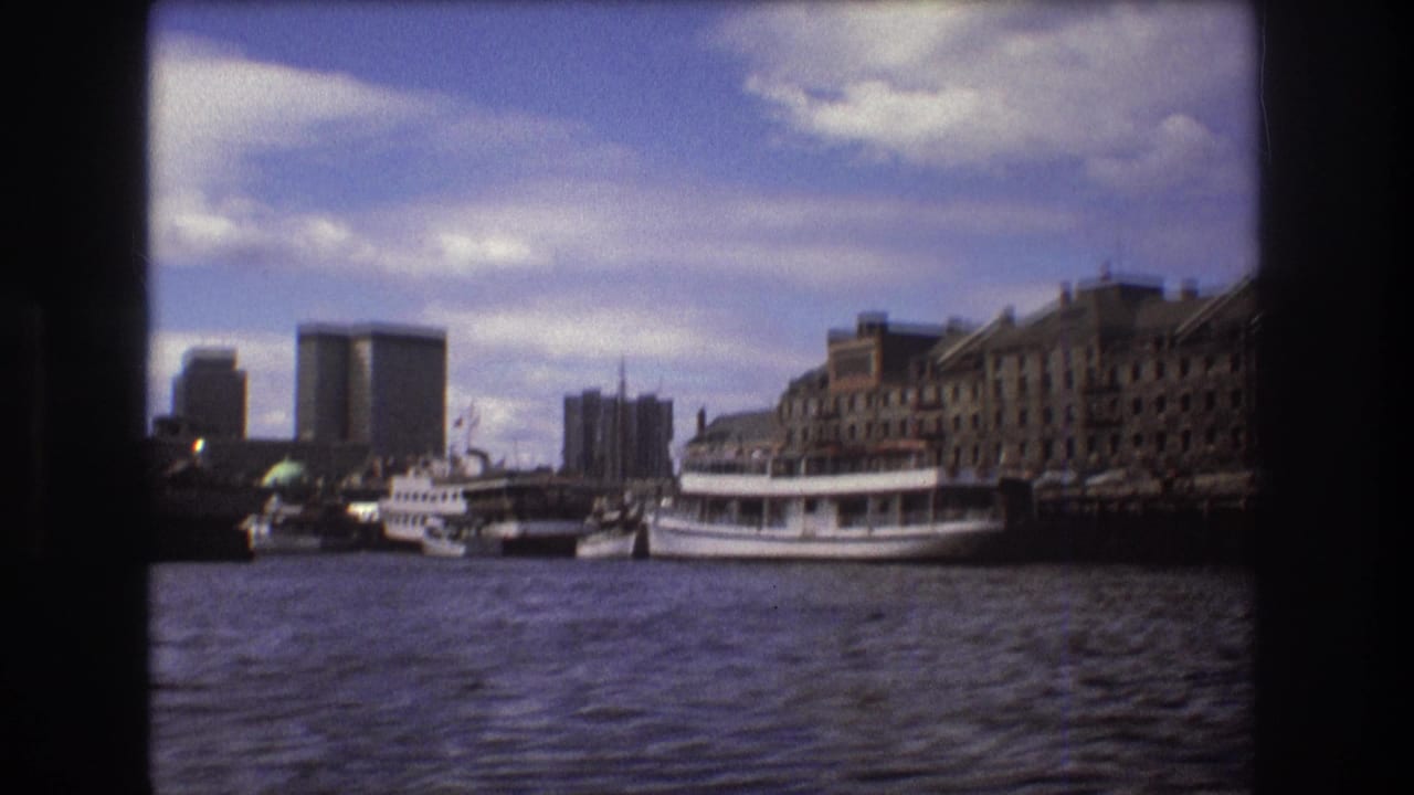 Boston waterfront seen from the harbor, with several docked white vessels, a larger passenger boat, mixed-height buildings, and a partly cloudy sky.