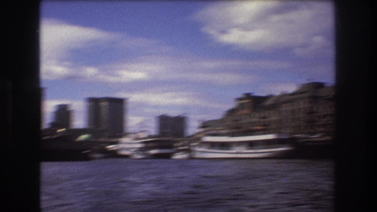 Motion-blurred 1975 Boston harbor frame from a moving boat, showing water, docked vessels, waterfront buildings, and distant high-rises.