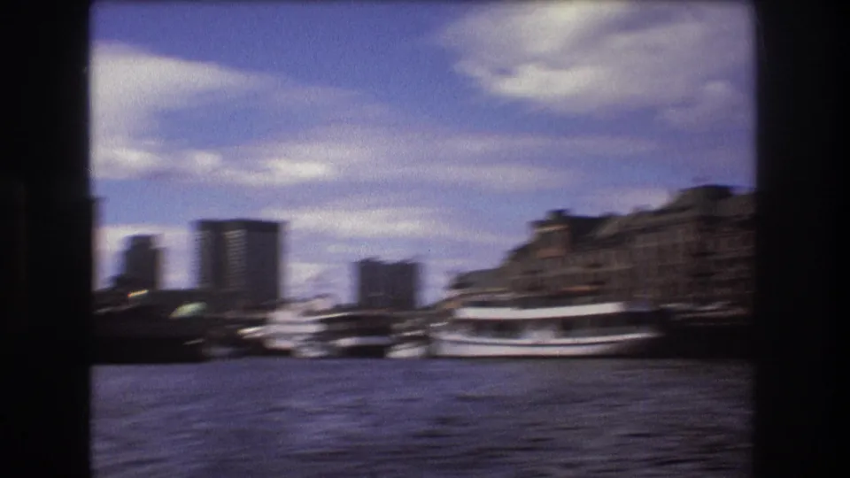 Motion-blurred 1975 Boston harbor frame from a moving boat, showing water, docked vessels, waterfront buildings, and distant high-rises.