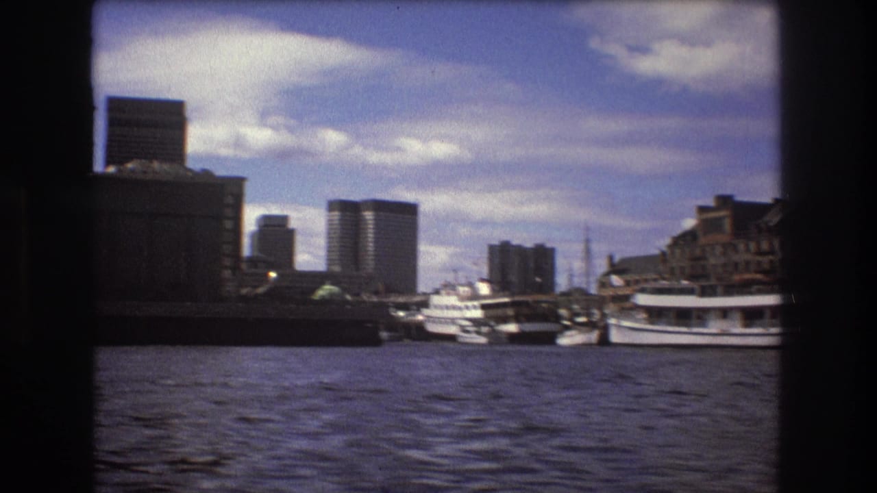 1975 Boston harbor view with rippling water in the foreground, docked boats at right, waterfront structures, and modern high-rise buildings beyond.