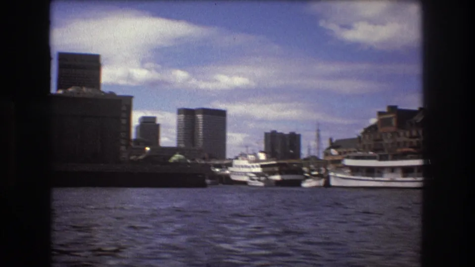 1975 Boston harbor view with rippling water in the foreground, docked boats at right, waterfront structures, and modern high-rise buildings beyond.