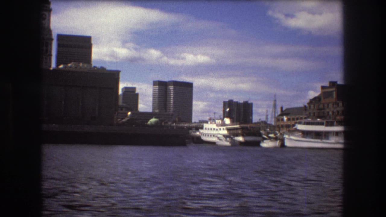 1975 Boston harbor view from a moving boat, showing water, moored boats, waterfront structures, and distant high-rise buildings beneath scattered clouds.