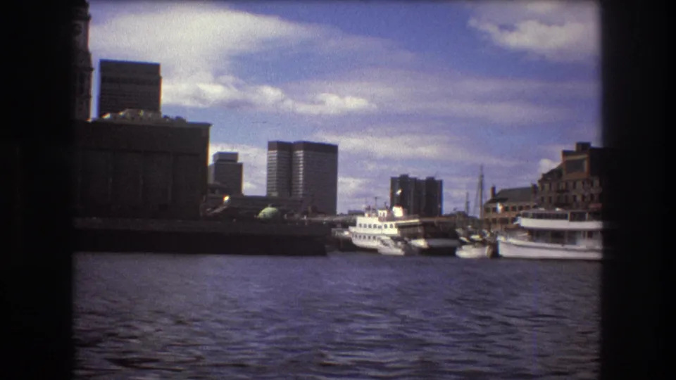 1975 Boston harbor view from a moving boat, showing water, moored boats, waterfront structures, and distant high-rise buildings beneath scattered clouds.