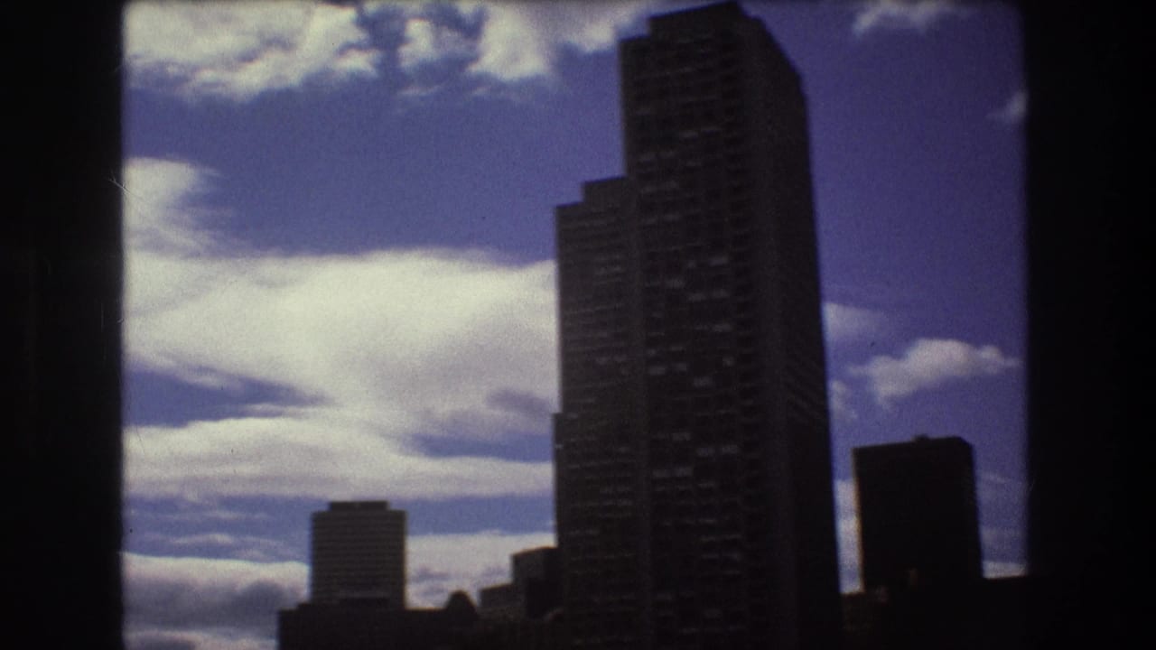 1975 Boston skyline detail with a dark high-rise tower and smaller buildings silhouetted against a blue sky filled with broad white clouds.
