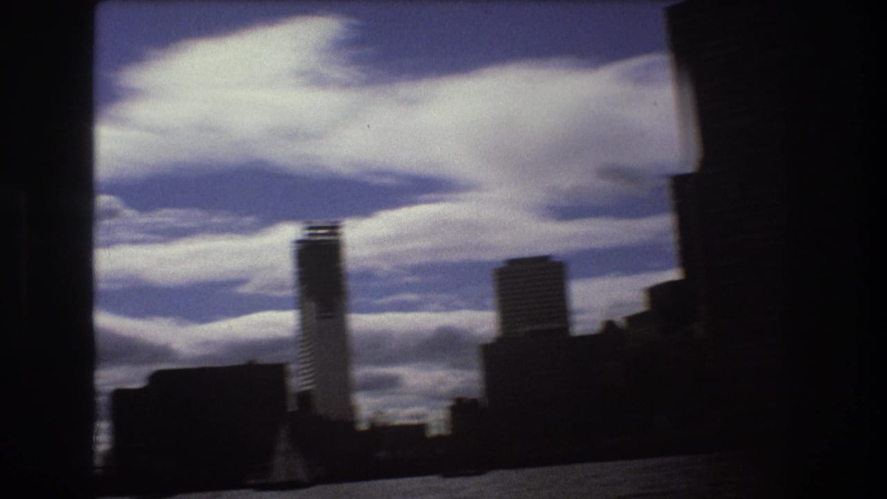 Boston harbor view from the water, showing silhouetted waterfront buildings and a tall tower against a blue sky with layered white clouds.