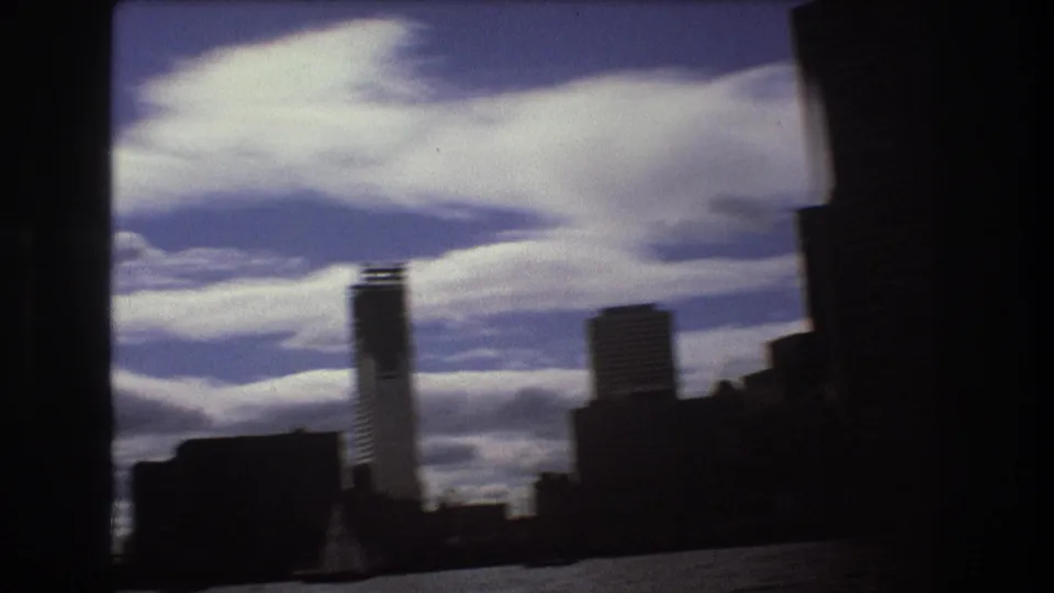 Boston harbor view from the water, showing silhouetted waterfront buildings and a tall tower against a blue sky with layered white clouds.