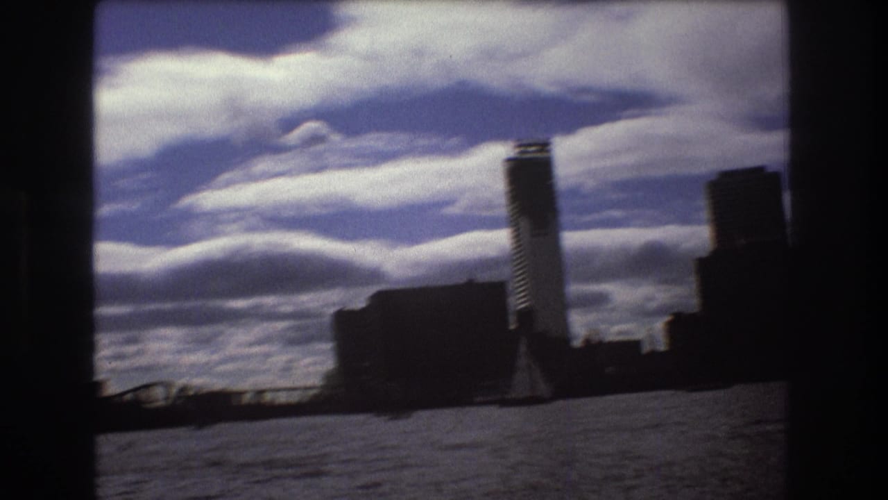 1975 Boston harbor home-movie frame with skyline silhouettes, rippled water, sweeping clouds, and a small sailboat near the lower left edge.