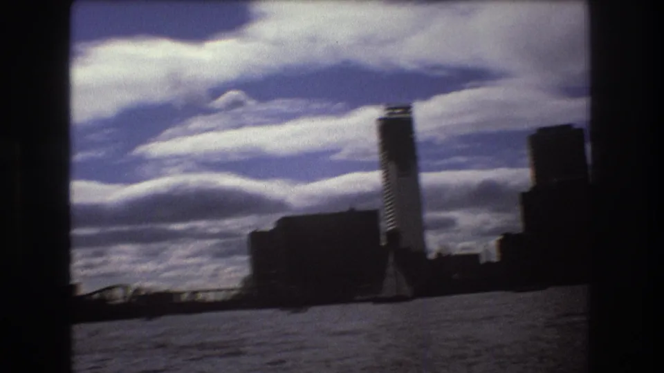 1975 Boston harbor home-movie frame with skyline silhouettes, rippled water, sweeping clouds, and a small sailboat near the lower left edge.