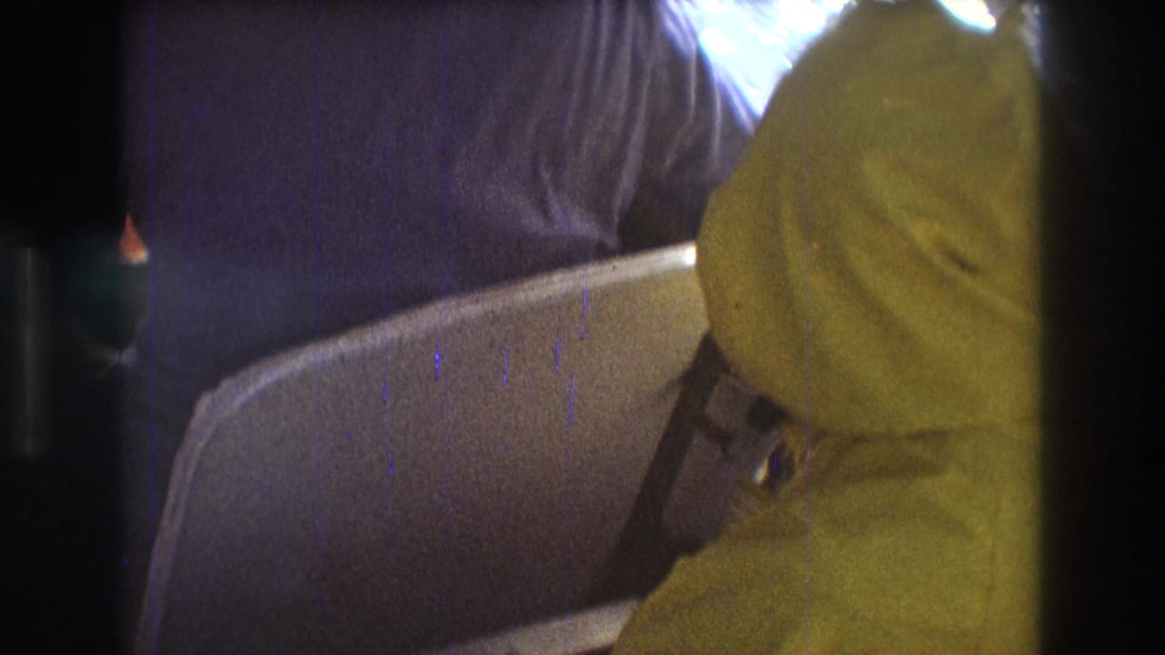 1975 Boston-area 8mm frame showing a passenger in a yellow hooded raincoat seated in a boat, with another figure partly visible and water outside the window.