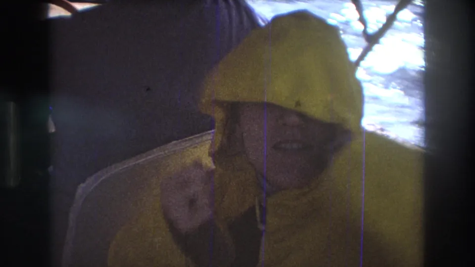1975 Boston-area 8mm frame of a person in a yellow hooded raincoat looking toward the camera while seated inside a boat with bright water beyond the window.