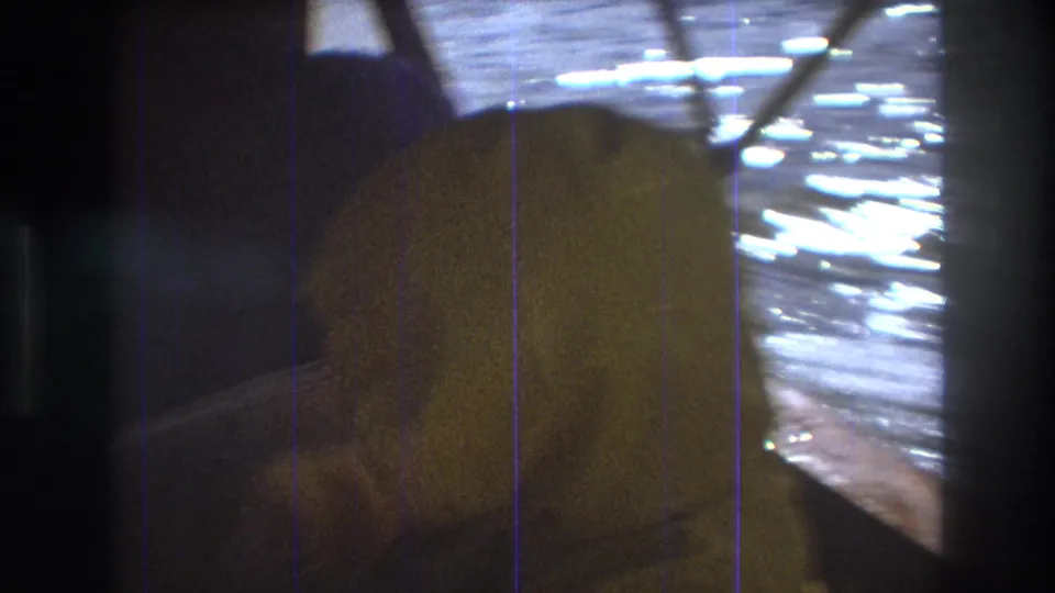 1975 Boston-area 8mm frame showing the back of a passenger in a yellow raincoat inside the boat, with reflective water and window framing in the background.