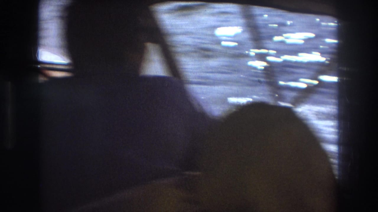 1975 Boston-area 8mm frame with a dark-shirted person partly silhouetted inside the boat as a yellow hooded passenger sits low in the foreground and sunlight reflects on the water outside.