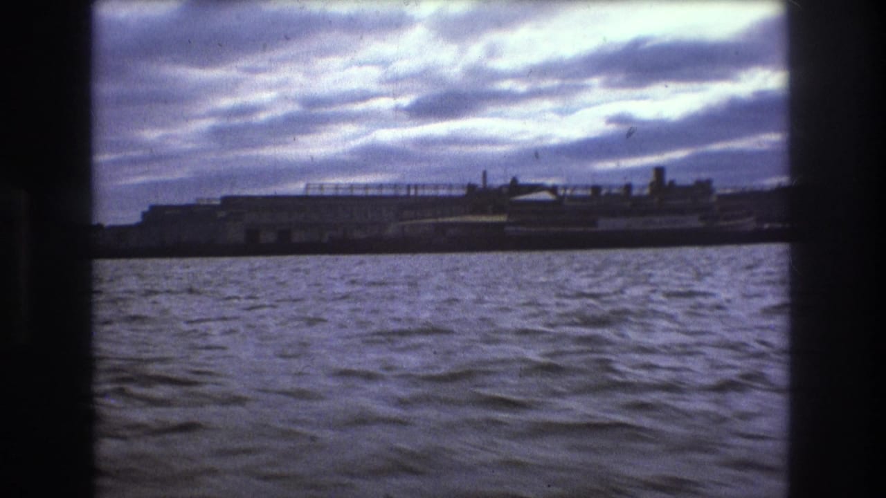 Boston 1975 8mm frame with a low industrial-looking waterfront and pier-like structures seen across rippled harbor water from a moving boat.