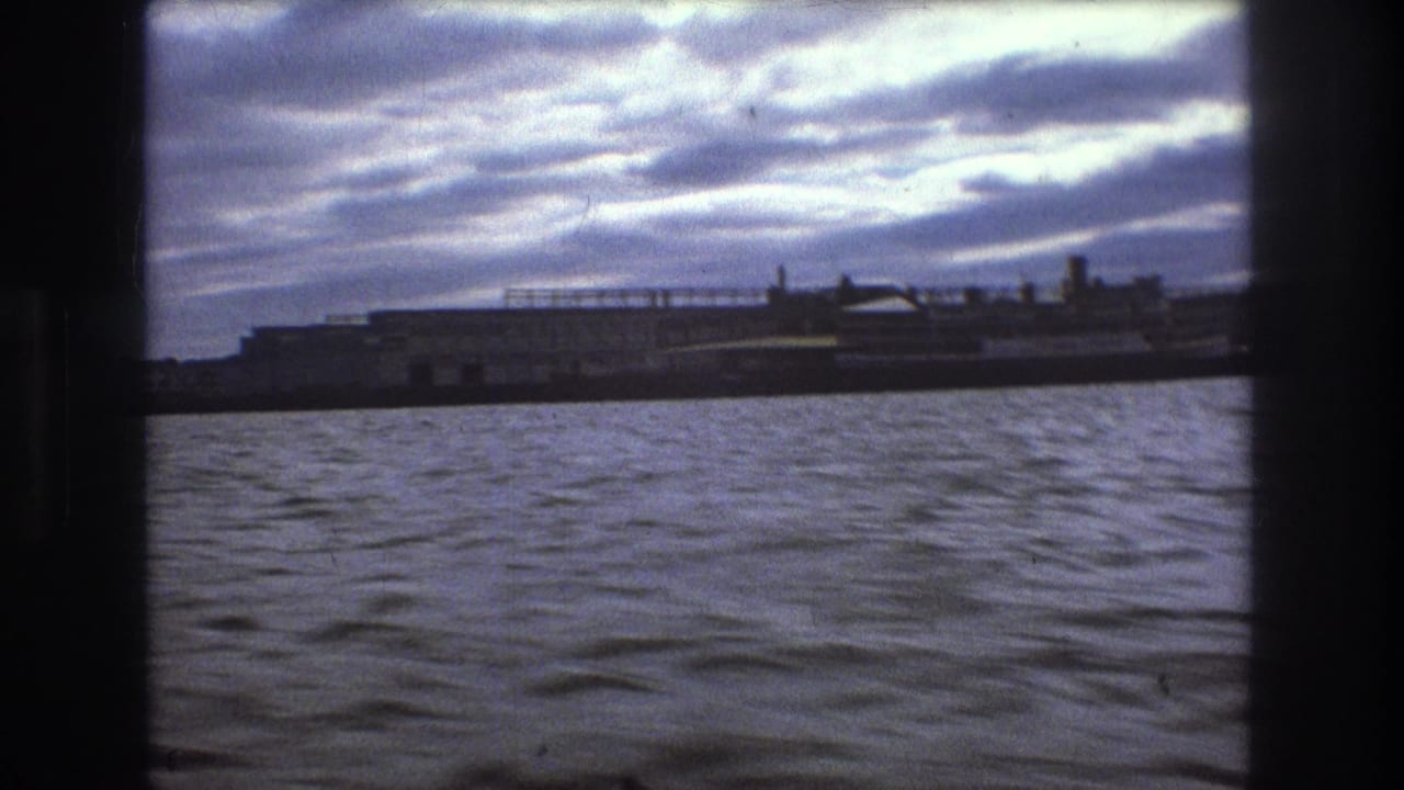 Boston 1975 8mm boat POV showing rippled harbor water, a dark shoreline strip, and distant waterfront structures beneath a bright cloudy sky.