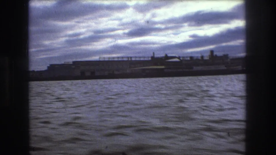Boston 1975 8mm boat POV showing rippled harbor water, a dark shoreline strip, and distant waterfront structures beneath a bright cloudy sky.