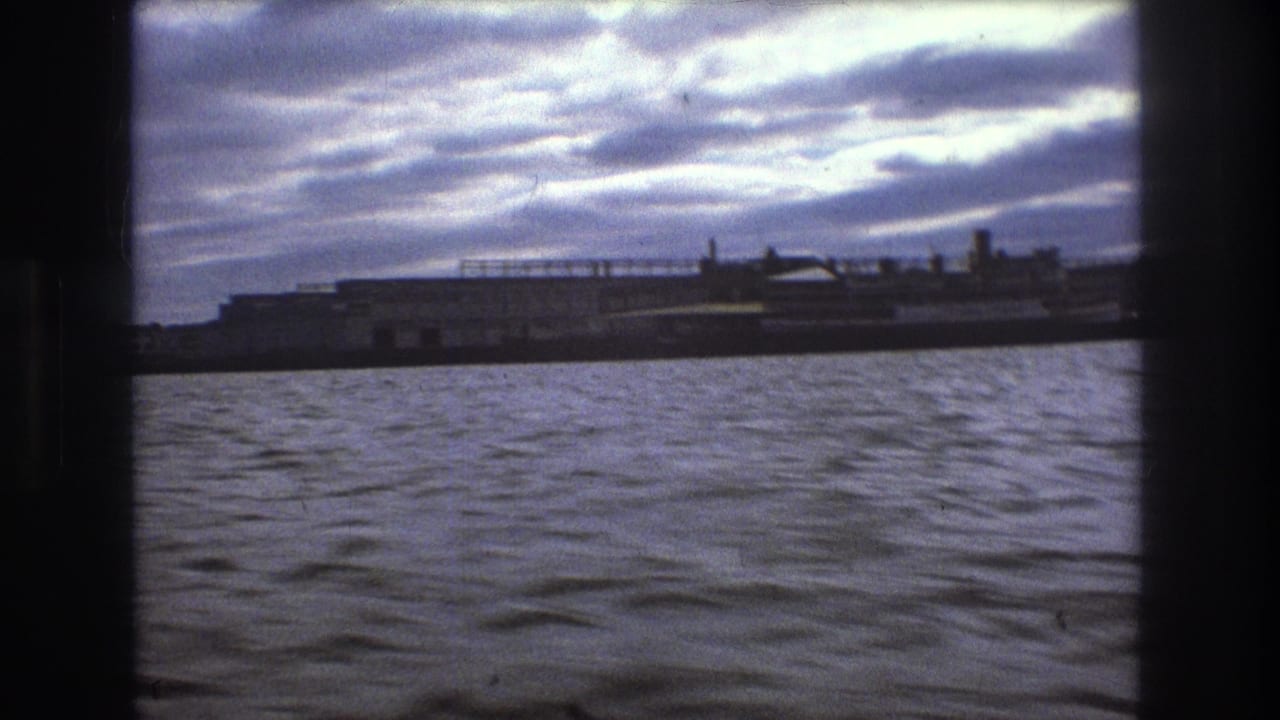 1975 Boston home-movie view from the water toward a low industrial waterfront or ship-like structure beneath layered clouds.