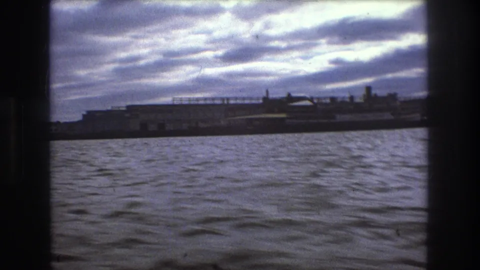 1975 Boston home-movie view from the water toward a low industrial waterfront or ship-like structure beneath layered clouds.