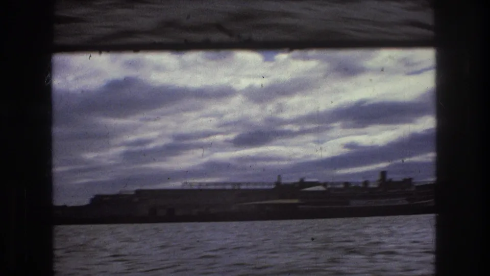 Boston-area harbor view from a moving vessel, with choppy water, low industrial forms, and a dramatic clouded sky in faded 8mm color.