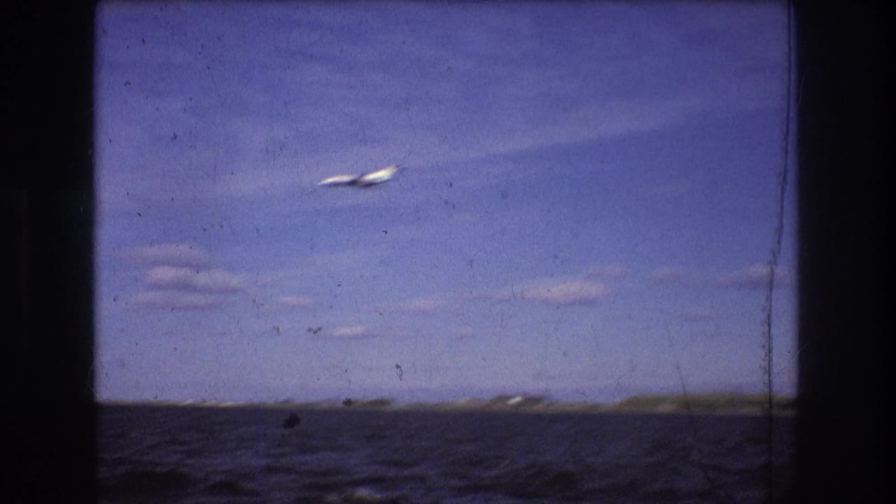 Wide 1975 Boston harbor home-movie frame with a small airplane crossing open sky above water and a distant low shoreline.