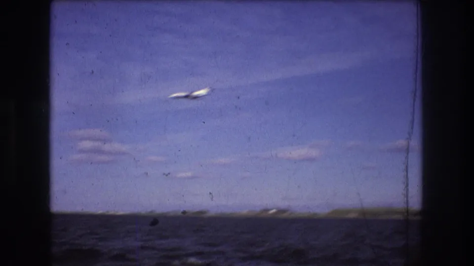 Wide 1975 Boston harbor home-movie frame with a small airplane crossing open sky above water and a distant low shoreline.