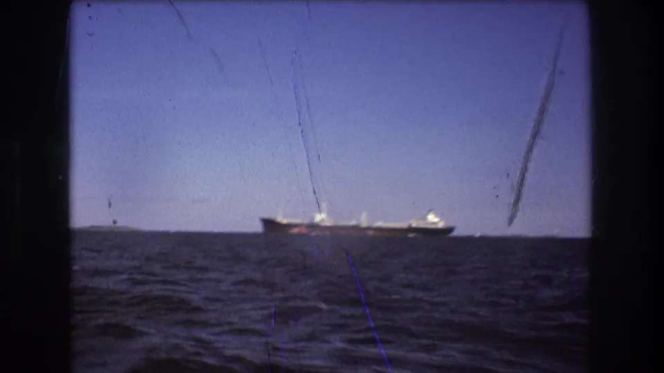1975 Boston-area 8mm frame with a large ship traveling near the horizon, seen across choppy water through a dark boat-window opening.