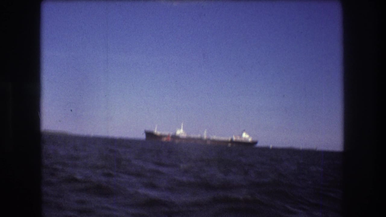 1975 Boston-area 8mm frame showing a large distant ship over blue water, with the horizon and shoreline softened by handheld home-movie focus.
