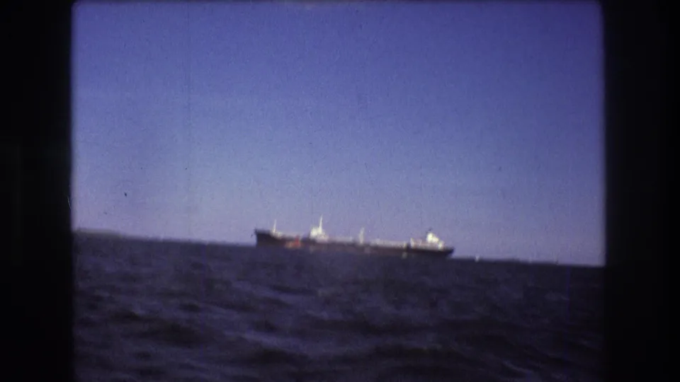 1975 Boston-area 8mm frame showing a large distant ship over blue water, with the horizon and shoreline softened by handheld home-movie focus.