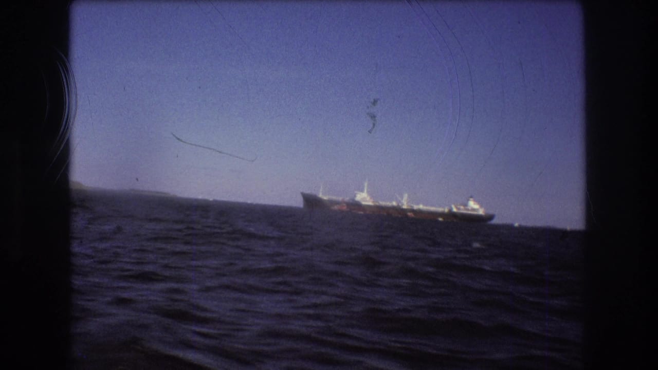 1975 Boston-area 8mm frame showing a large ship near low distant land, filmed from a moving boat across dark blue choppy water.