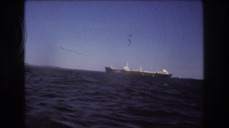 1975 Boston-area 8mm frame showing a large ship near low distant land, filmed from a moving boat across dark blue choppy water.