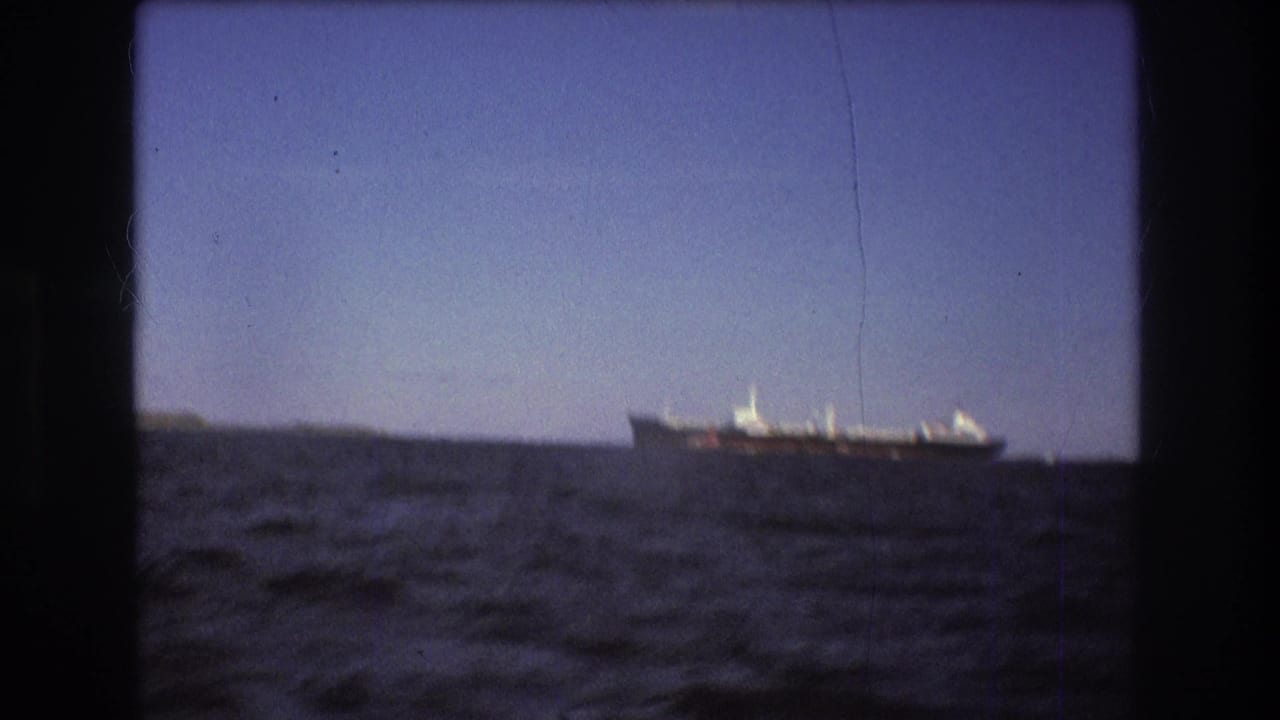 Boston-area 1975 8mm frame showing a distant large commercial ship on open water, with dark film borders and a low horizon.