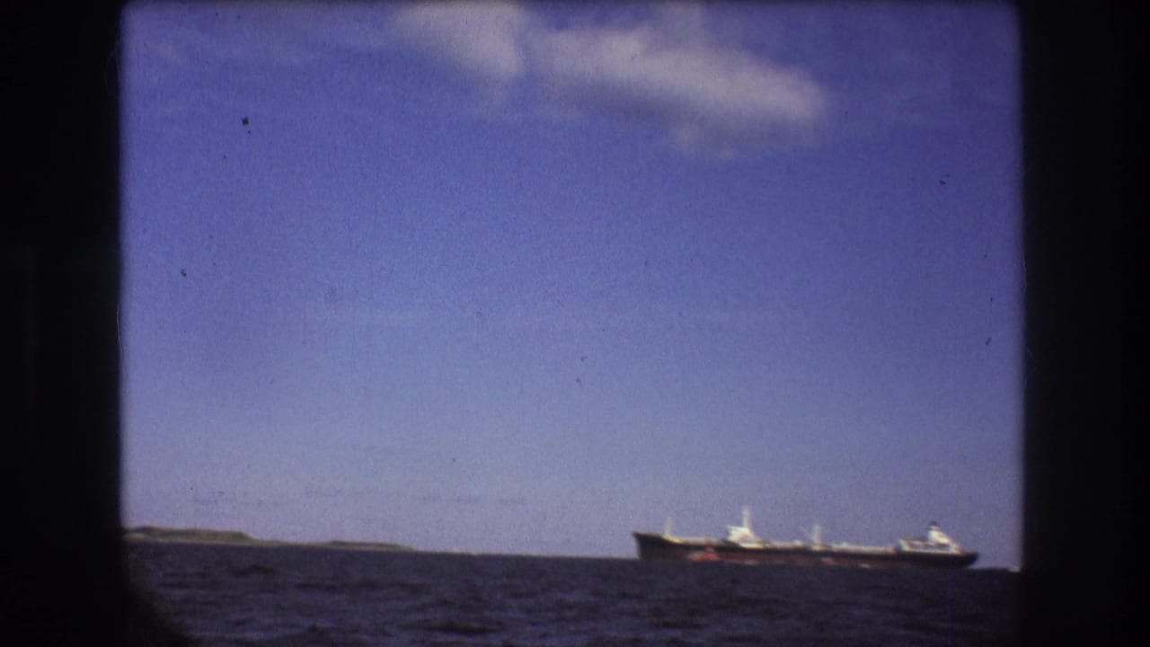 A large commercial vessel sits near the horizon in 1975 Boston-area home-movie footage, framed by blue sky, dark sea, and visible 8mm gate edges.
