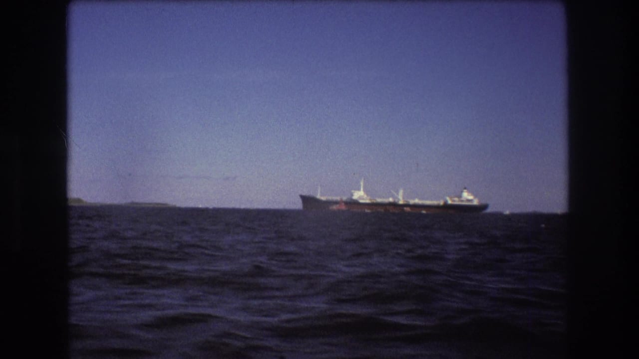 Distant commercial ship filmed from a moving boat near Boston in 1975, with rippling foreground water and faint low shoreline behind it.