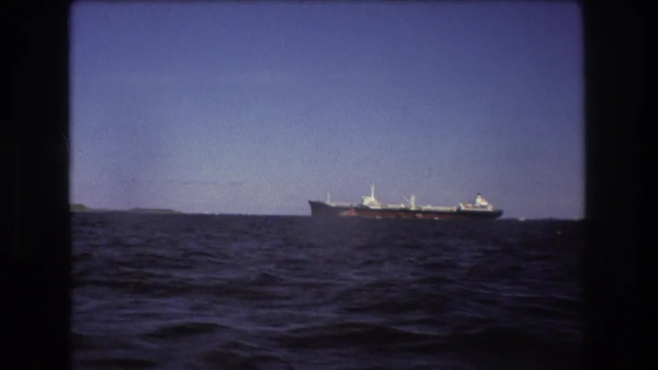 Distant commercial ship filmed from a moving boat near Boston in 1975, with rippling foreground water and faint low shoreline behind it.