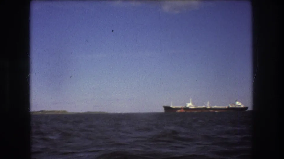 Final 8mm frame of the Boston-area ship view, showing the vessel at the right edge of the horizon beyond open water and a small stretch of land.
